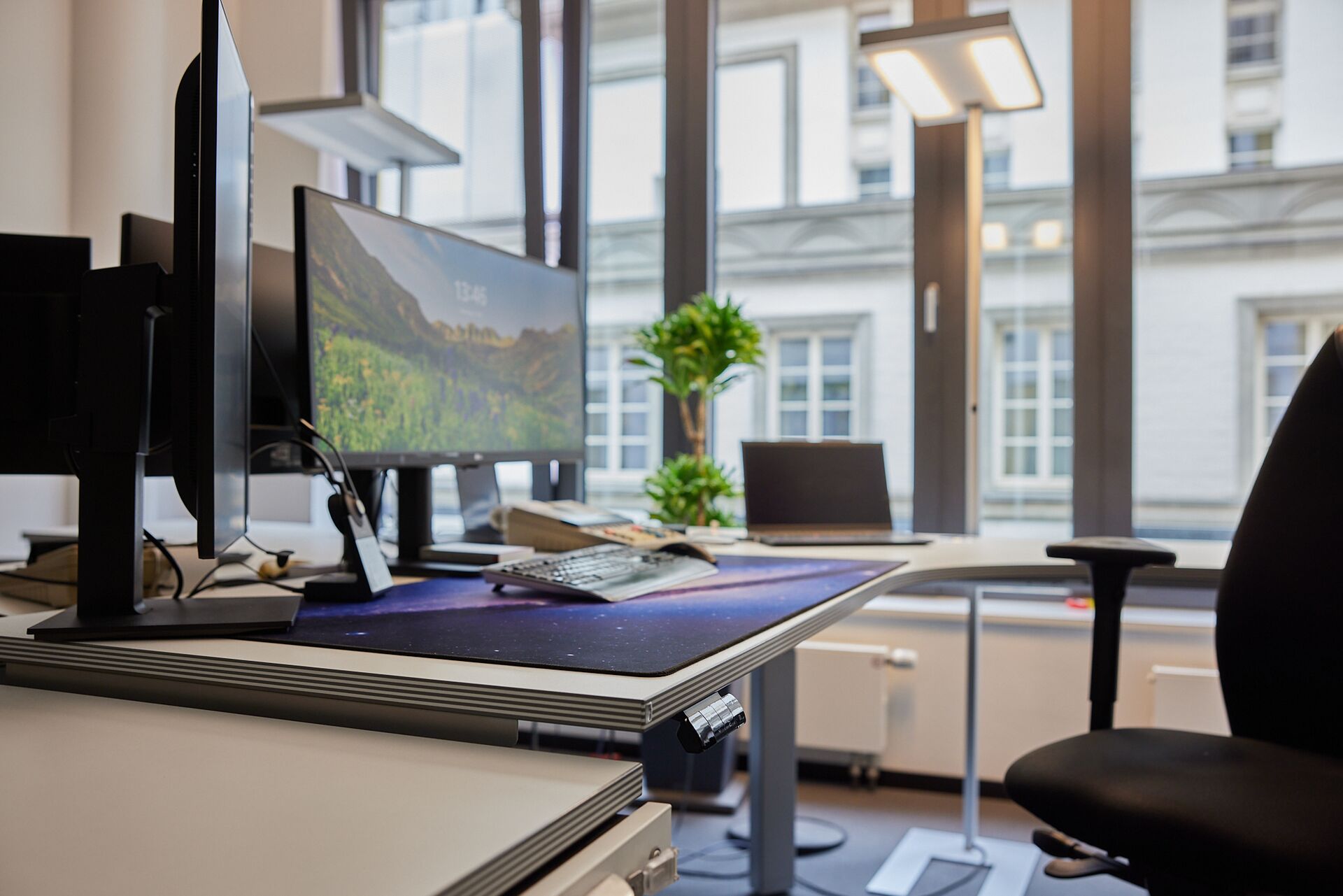 Nahaufnahme eines modernen Arbeitsplatzes mit zwei Monitoren, höhenverstellbarem Schreibtisch und Blick auf große Fenster mit Stadtansicht.
